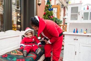 Noel aile tatili konsepti. Mutlu Noeller ve mutlu tatiller. Anne ve kızı Noel Baba kostümü giymiş. Eğlenceli yürüyüşler. Anne kızını kollarında tutuyor, samimi bir gülümseme..