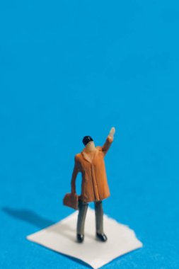 Turuncu bir ceket giymiş küçük bir heykel bir elinde evrak çantası tutarken bir eliyle el sallıyor. Arka plan parlak bir mavidir, neşeli bir sahne yaratır..