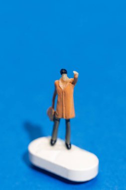 Bir elinde evrak çantası tutarken bir elinde turuncu bir ceket giymiş küçük bir heykel dalgalanır. Beyaz bir zeminde duruyor, parlak mavi bir arka plana karşı..