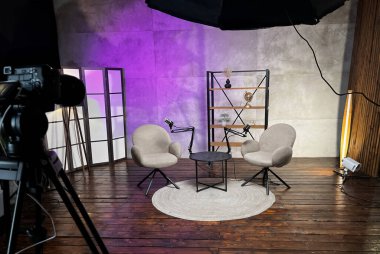 Modern bir podcast stüdyosunda iki rahat sandalye ve kayıt için hazır küçük bir masa yer alır. Yumuşak ışıklandırma ve dekoratif zemin tartışmalar için davetkar bir ortam yaratır..