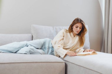 Evindeki yumuşak bir kanepede uzanmış, kitabına dalmış bir kız. Zamanını çalışarak ve derslerinden zevk alarak geçirdikçe rahatlamış görünüyor..