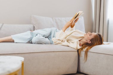 Bir kız yumuşak bir kanepede uzanıyor, rahat evinde kitabın tadını çıkarıyor. Rahatlamış, rahat giyinmiş ve huzurlu bir ortamda okumaya odaklanmış..