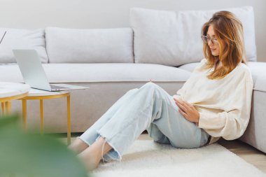 Serbest çalışan bir öğrenci yerde oturuyor, günlük kıyafetler giyiyor, dizüstü bilgisayarda çalışıyor. Sakin atmosferde rahat bir yaşam alanı var. Yumuşak ışıklandırma ve rahat bir halı..