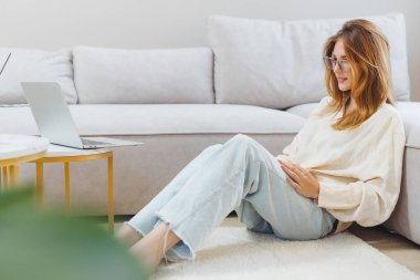 Bir öğrenci online çalışma için bir dizüstü bilgisayar kullanarak evde yerde rahatça oturur. Konforlu ortamda yumuşak bir halı ve açık renkli bir kanepe var. Rahat bir atmosfer yaratıyor..