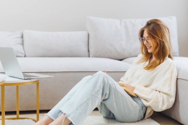 Genç bir kadın oturma odasında yumuşak bir halının üzerinde oturuyor, serbest çalışmalarıyla meşgul. Açık dizüstü bilgisayarının yanında notlar alırken gülümsüyor, rahat atmosferin tadını çıkarıyor..