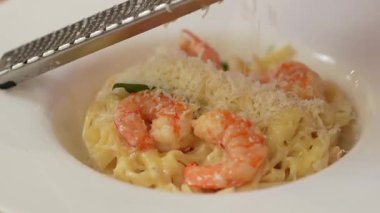 Bir şef, kremalı soslu, taze otlu, lezzetli bir karides makarna yemeği hazırlar ve bol miktarda rendelenmiş peynir, sıcak bir mutfakta aşçılık becerileri sunar..