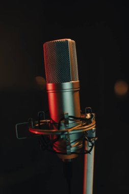 Kayıt stüdyosunda bir mikrofon duruyor. Stüdyonun ışığı az ve renkli ışıklar mikrofon gövdesine yansıyor. Bu ayar ses kaydına odaklanmayı gösterir.