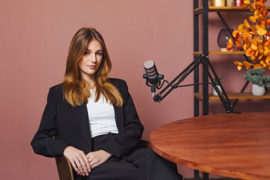 Bir kadın stüdyoda ahşap bir masada oturuyor. Podcast röportajı için hazırlanıyor. Önüne ayarlanabilir bir kolda bir mikrofon yerleştirildi. Arkaplan dekorasyonu gösterir.