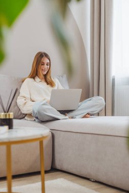 Oturma odasındaki kanepede oturan bir kadın dizüstü bilgisayarında yazıyor. İşine odaklanmış ve pencereden gelen yumuşak, doğal ışıkla çevrili..