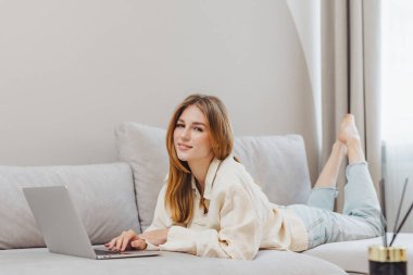 Serbest çalışan bir öğrenci evde dizüstü bilgisayar kullanıyor. Ayakları havada bir kanepede uzanıyor, derslerine odaklanıyor. Yumuşak ışık pencereden girer.