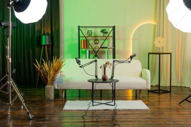 Bir podcast stüdyosunda bir masada iki mikrofon bulunur. Arka planda bir kanepe, bitkiler ve ışıklar var. Tartışmaları kaydetmek ya da röportajları yapmak için ekipman ayarlandı.