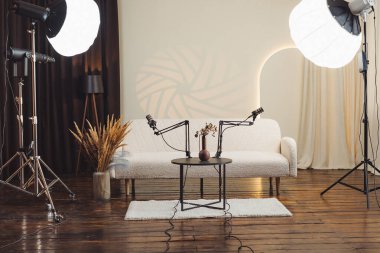 Bir podcast stüdyosunda kollara monte edilmiş iki mikrofon, vazolu küçük bir masa ve yumuşak ışıklandırma yer alır. Bölgede bir kanepe ve ahşap döşeme var. Alan, tartışmaları kaydetmek için hazır..