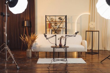 Podcast stüdyosundaki bir masaya iki mikrofon yerleştirildi. Arka planda bir kanepe ve dekoratif eşyalar var. Yumuşak ışıklandırma kayıt için bir karşılama alanı yaratır.