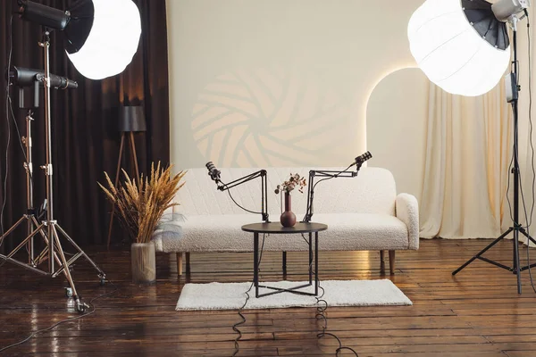 Bir podcast stüdyosunda kollara monte edilmiş iki mikrofon, vazolu küçük bir masa ve yumuşak ışıklandırma yer alır. Bölgede bir kanepe ve ahşap döşeme var. Alan, tartışmaları kaydetmek için hazır..