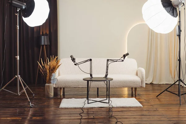 Podcast stüdyosundaki küçük bir masaya bakan iki mikrofon var. Yumuşak ışıklar mekanı aydınlatarak kayıt için davetkar bir atmosfer yaratıyor. Yakınlarda bir kanepe var..