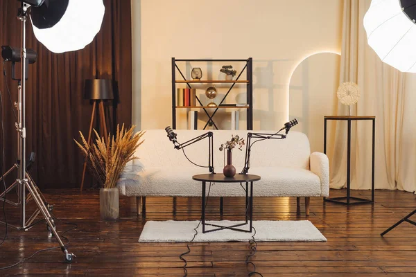 Podcast stüdyosundaki bir masaya iki mikrofon yerleştirildi. Arka planda bir kanepe ve dekoratif eşyalar var. Yumuşak ışıklandırma kayıt için bir karşılama alanı yaratır.