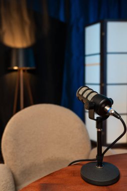 Podcast stüdyosundaki bir masanın üzerinde bir mikrofon duruyor. Yakınlarda bir sandalye var. Yumuşak ışıklandırma rahat bir atmosfer yaratır. Arka planda bir disk bölümü kurulumu tamamlıyor.