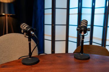 Podcast stüdyosundaki ahşap bir masada iki mikrofon duruyor. Oda kayıt için ayarlandı, mavi ve siyah bir arkaplanı var. Sandalyeler, ev sahiplerinin oturup konuşması için yerleştirildi..