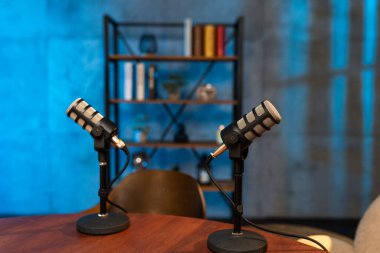 İki mikrofon stüdyo ortamında bir masanın üzerinde duruyor. Kitaplar ve dekorasyonlar onların arkasındaki raflarda görülebilir. Podcaster 'lar bir sonraki bölümlerini kaydetmeye hazır olun..