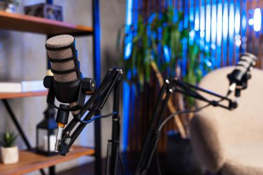 Podcast kaydı için iki mikrofon kuruldu. Arkaplanda bitkilerle dolu raflar ve rahat bir oturma alanı yer alıyor. Yumuşak ışıklandırma sohbet için rahat bir atmosfer yaratır..