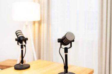 Podcast kaydı için stüdyodaki ahşap bir masada iki mikrofon duruyor. Arka planda yumuşak bir lamba ve ışık perdeleri var. Atmosfer röportaj için hazır..