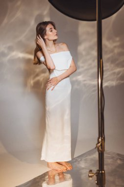 Beyaz bir elbise giymiş bir kız moda stüdyosunda duruyor. Stüdyo ışıklarının yanında poz veriyor. Işık duvarda yumuşak desenler oluşturuyor..