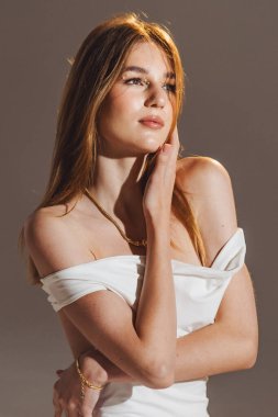 Beyaz elbiseli bir kız fotoğraf stüdyosunda duruyor. O, yüzüne dokunurken ve doğrudan kameraya bakarken güven içinde poz verir. Işıklandırma onun özelliklerini açıkça vurguluyor..