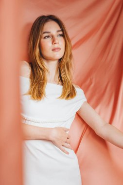 Beyaz elbiseli bir kız fotoğraf stüdyosunda poz veriyor. Yumuşak perdelerin yanında duruyor ve düşünceli bir şekilde kameraya bakıyor. Ayar parlak ve onun varlığına odaklı..