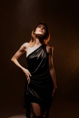 Genç bir model, parlak siyah bir elbise giyerek bir fotoğraf stüdyosunda güvenle duruyor. Yumuşak ışıklandırma onun zarif duruşunu vurguluyor. Sahne bir moda ve sanatsal anı yakalar..