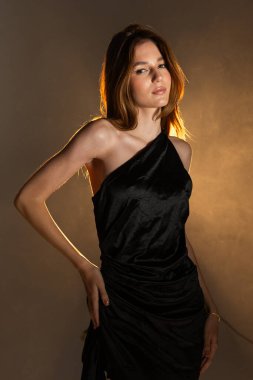 Bir kız sıcak ışıklandırması olan bir stüdyoda güven içinde duruyor. O, pozunu vurgulayan siyah bir elbise giyer. Sade bir arkaplan ile ayarlar basittir.