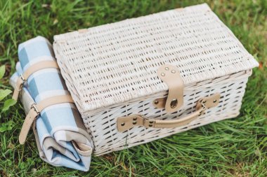 Piknik sepeti yeşil çimlerin üzerinde rulo battaniyenin yanında duruyor. Sahne, insanların dışarıda yemek yediği güneşli bir öğleden sonrayı gösteriyor..