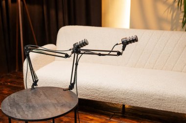 Stüdyoda bir masada iki mikrofon standı var. Arka planda açık renkli bir kanepe görülebilir. Burası podcast kayıt oturumları için bir yerdir.