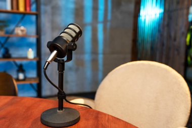 Modern bir odada rafları eşyayla dolu bir masanın üzerinde bir mikrofon duruyor. Masanın yanına bir sandalye yerleştirildi, yakında bir podcast yapılacağını gösteriyor..