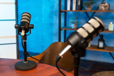 Podcast kaydı için tasarlanmış bir stüdyoda tahta bir masanın üzerinde iki mikrofon duruyor. Mavi arka plan modern bir dokunuş katıyor. Kitaplar ve bir bitki rafta görünür..