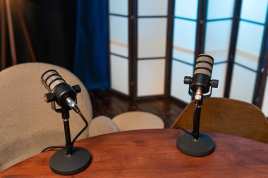 Kayıt alanındaki ahşap bir masada iki mikrofon oturuyor. Sandalyeler yakında. Yumuşak arka planlar podcast konuşmaları için bir yer öneriyor. Ekipman kayıt için hazır..