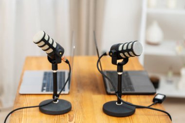 Tahta bir masada iki mikrofon duruyor. Her mikrofon bir dizüstü bilgisayara bağlı. Kurulum bir podcast kayıt oturumu için. Odada hafif ışıklandırma var..
