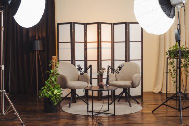 Podcast için stüdyoda iki sandalye ve bir masa hazırlanıyor. Masanın üzerine mikrofonlar yerleştirildi ve stüdyo ışıkları bölgeyi çevreledi. Bitkiler çevreye eklenir.