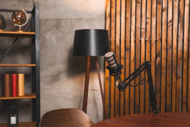 Podcast stüdyosundaki bir masanın üzerine bir mikrofon yerleştirildi. Yakınlarda bir lamba duruyor. Raflar kitap ve dekor tutuyor, kayıt için çalışma alanı yaratıyor.