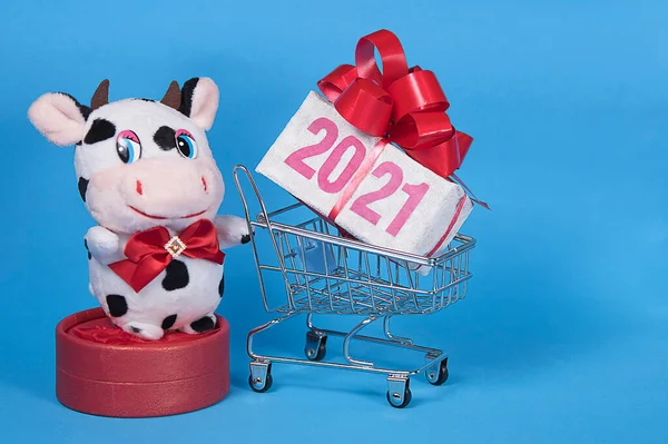 Happy Christmas bull or cow toy and shopping trolley with 2021 text on gift box. Blue background. Greeting card. Sale concept.