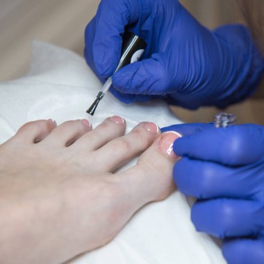 The pedicurist covers the nails with pink varnish. Hands in blue gloves cares about a woman`s foot nails.	
