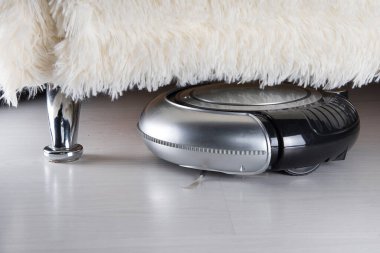 Robot vacuuming under sofa. Black and silver robotic vacuum cleaner on white laminate wooden floor.  clipart