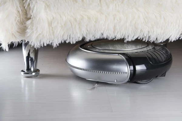 Robot kanepenin altını süpürüyor. Beyaz lamine ahşap zeminde siyah ve gümüş robotik elektrikli süpürge.. 