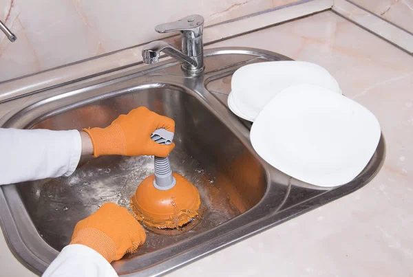 Woman's hands with orange gloves cleaning sewer and water at kitchen faucet over metal sink. Female hand with plunger. Home service concept