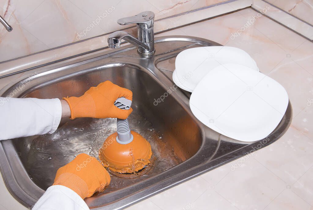 Woman's hands with orange gloves cleaning sewer and water at kitchen faucet over metal sink. Female hand with plunger. Home service concept