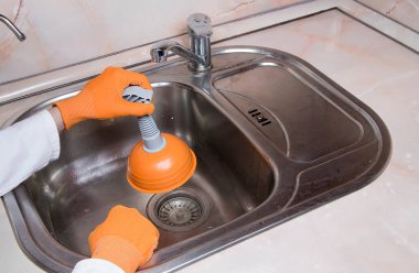 Woman's hands with orange gloves cleaning sewer at kitchen faucet over metal sink. Close up of hand with plunger clipart