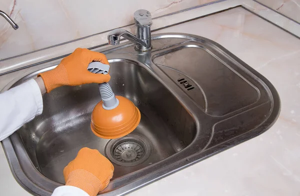 Woman\'s hands with orange gloves cleaning sewer at kitchen faucet over metal sink. Close up of hand with plunger