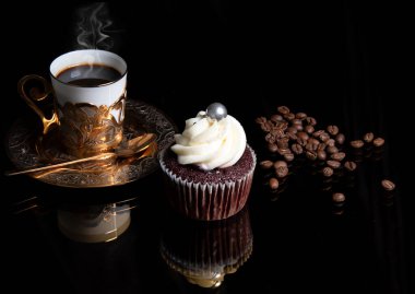 Buharlı altın kahve fincanı, ev yapımı kek ve siyah arka planda kahve çekirdekleri, kremalı ev yapımı kek. 