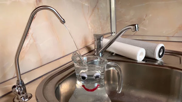 Pouring filtered water into happy glass pitcher from water filter. Closeup of jug, sink and faucet. Drinkable water in kitchen