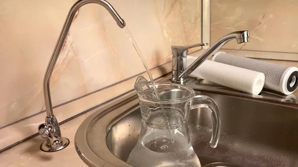 Pouring filtered water into glass pitcher from water filter. Closeup of jug, sink and faucet. Drinkable water in kitchen
