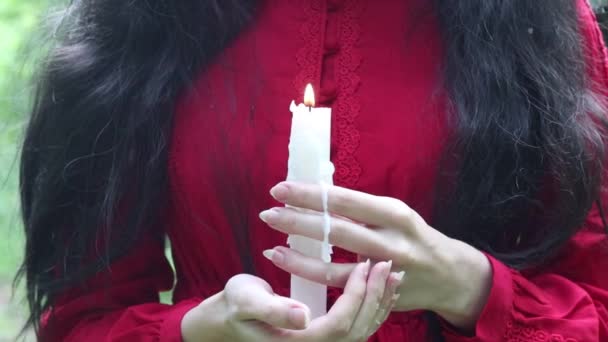 Дівчина в етнічній червоній сукні тримає палаючу воскову свічку в темному лісі . — стокове відео